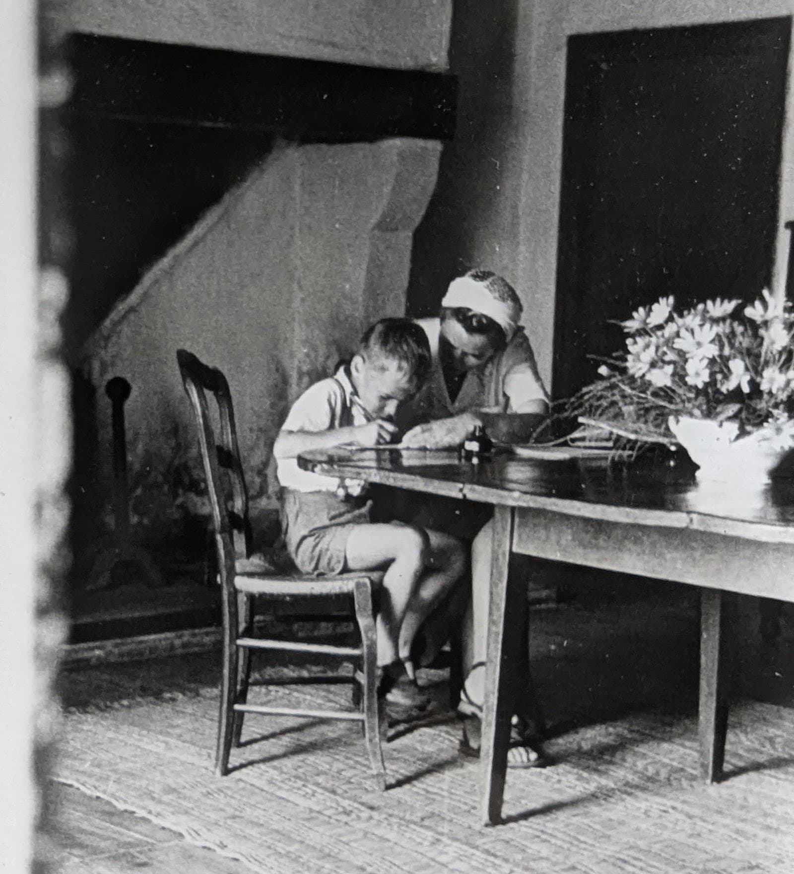 A black-and-white family photo from the 1950s: a child writing at a large wooden table, beside a woman.