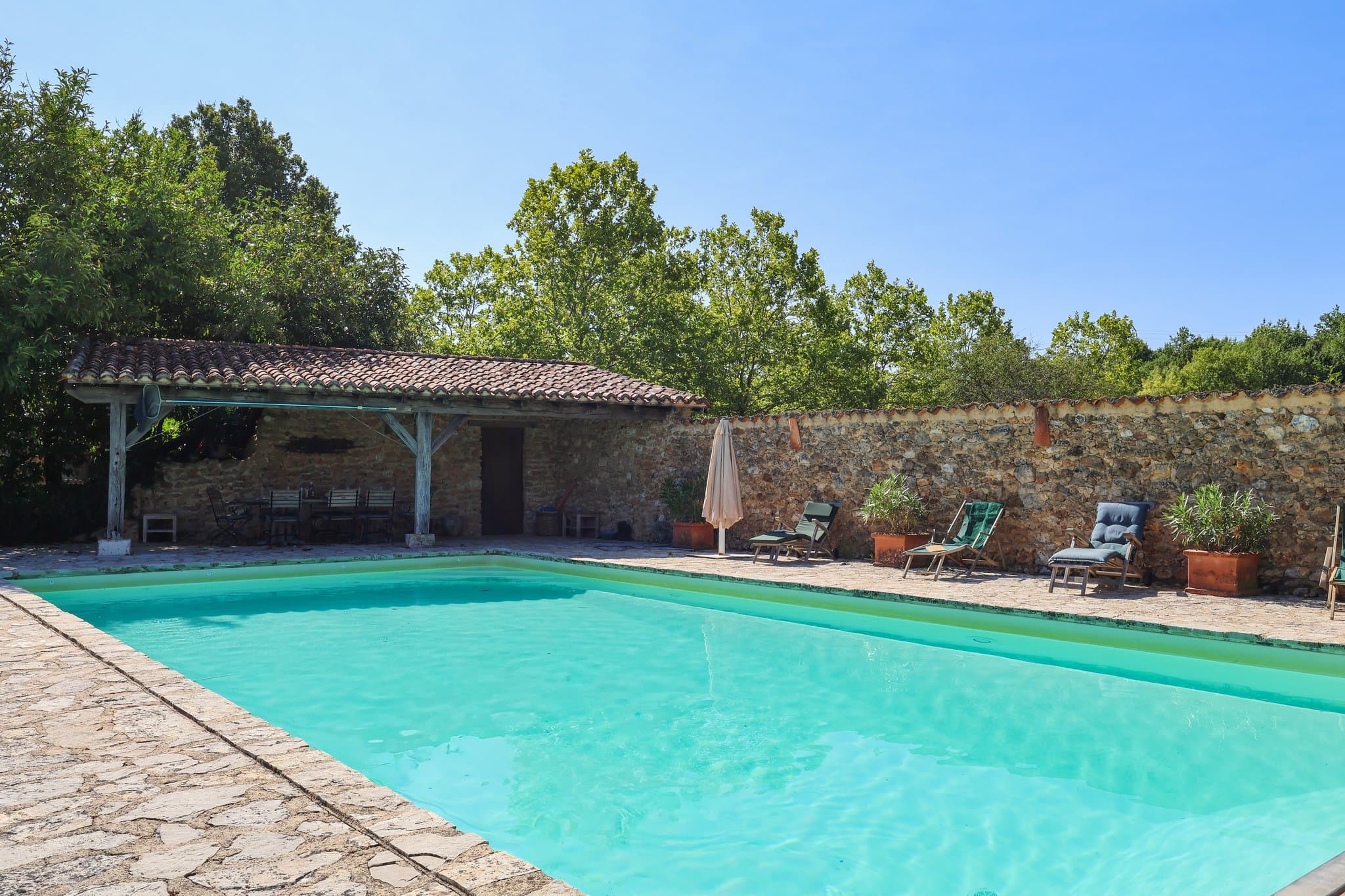 The pool at Le Clos de Moncouche, edged by a stone terrace: under the tiled roof of the covered terrace, a long table and its chairs; along the stone wall, deckchairs and oleanders in terracotta pots.